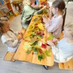 Dzieci przy drewnianym stole układają kolorowe liście. W tle widać regały z książkami. Przeważają odcienie zieleni, żółci i czerwieni.