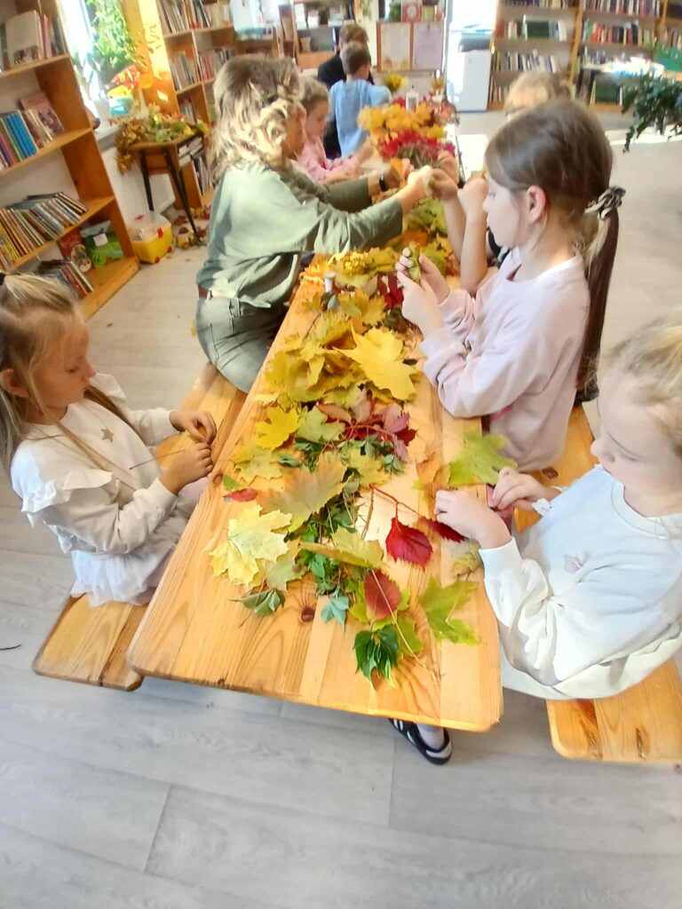 Dzieci przy drewnianym stole układają kolorowe liście. W tle widać regały z książkami. Przeważają odcienie zieleni, żółci i czerwieni.