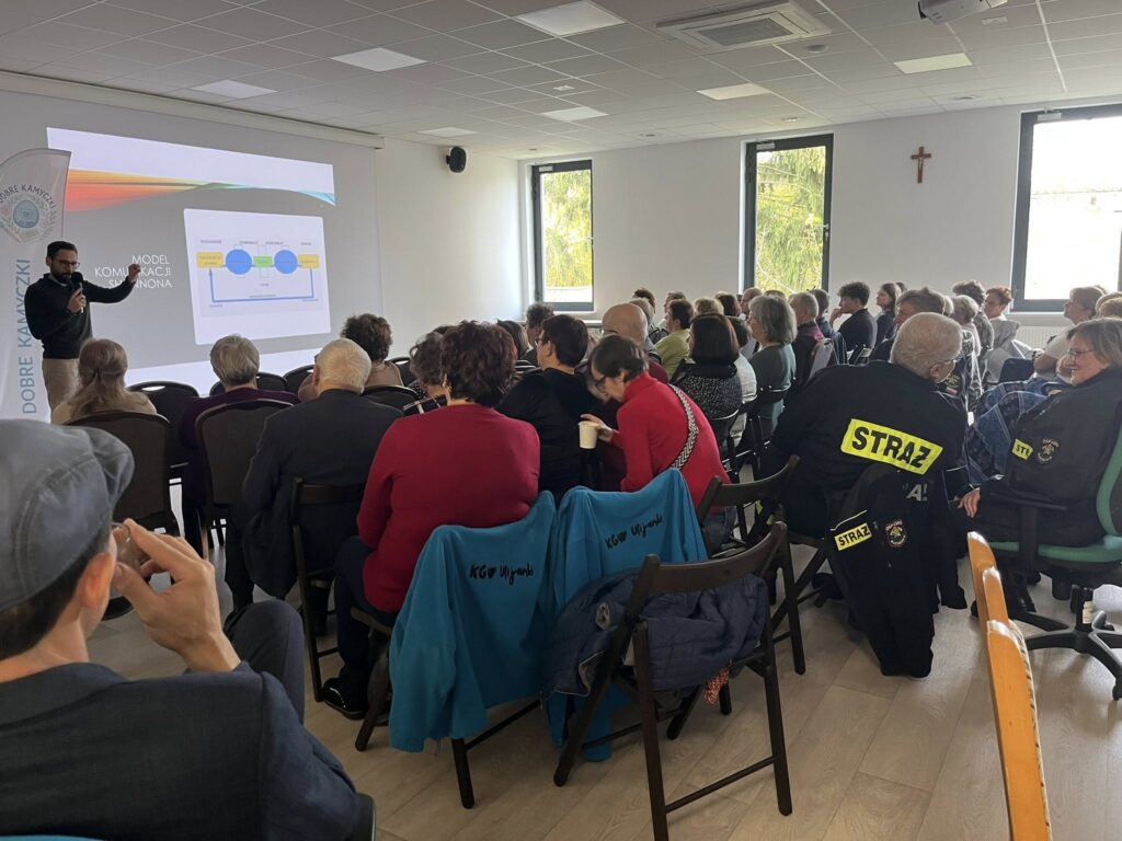 Spotkanie w jasnej sali, ludzie siedzą przed prezentacją na projektorze. Na ścianie czarny krzyż, dwie duże okna widoczne.