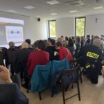 Spotkanie w jasnej sali, ludzie siedzą przed prezentacją na projektorze. Na ścianie czarny krzyż, dwie duże okna widoczne.