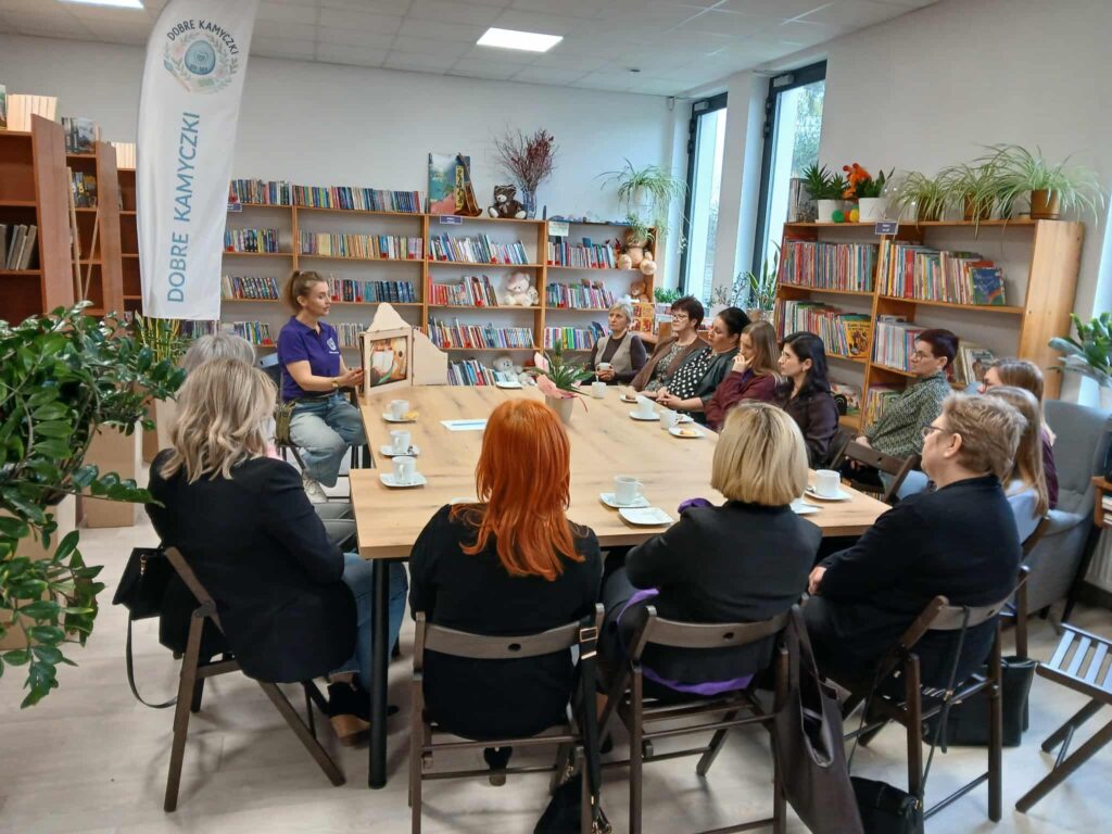 Na obrazie widać grupę ludzi siedzących wokół drewnianego stołu w bibliotece. Kobieta prowadzi prezentację, a w tle są regały z książkami. Ściany są jasne, z dużymi oknami i zielonymi roślinami.