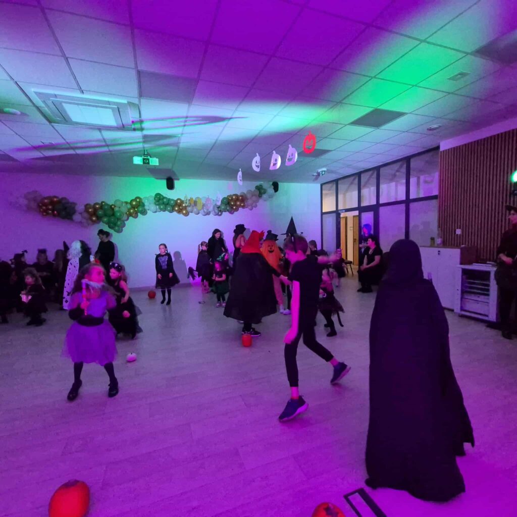 Dzieci w przebraniach na przyjęciu Halloween. Fioletowe i zielone oświetlenie. Ściany ozdobione balonami i dyniami.