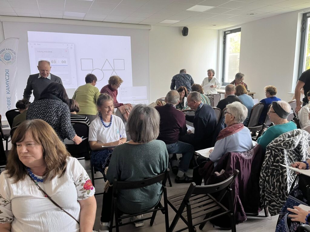 Grupa osób siedzi na czarnych krzesłach w sali z projektorem i jasnymi ścianami. W tle duże okna, na ekranie figury geometryczne. Uczestnicy ubrani są kolorowo, rozmieszczeni w grupach.