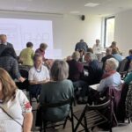 Grupa osób siedzi na czarnych krzesłach w sali z projektorem i jasnymi ścianami. W tle duże okna, na ekranie figury geometryczne. Uczestnicy ubrani są kolorowo, rozmieszczeni w grupach.