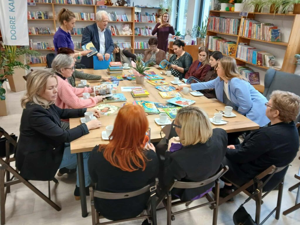 Obraz przedstawia grupę osób siedzących przy stole w bibliotece. Na stole leżą kolorowe książki. W tle widoczne są regały pełne książek i rośliny.