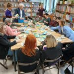 Obraz przedstawia grupę osób siedzących przy stole w bibliotece. Na stole leżą kolorowe książki. W tle widoczne są regały pełne książek i rośliny.