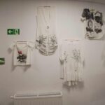 Obraz przedstawia wystawę białych bluzek z czarnymi i kolorowymi wzorami na ścianie. W tle biała ściana oraz zielony znak ewakuacyjny. Ubrania mają różne długości rękawów i detale graficzne.