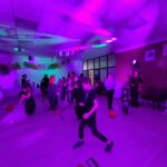 Na zdjęciu dzieci przebrane za postacie na Halloween bawią się w fioletowym świetle. W tle balony i ozdoby. Pomieszczenie jest przestronne i zaciemnione.