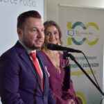 Mężczyzna w garniturze i kobieta w sukni stoją przy mikrofonie. Tło z plakatami w jasnych kolorach i napisami. Jasne oświetlenie na scenie.