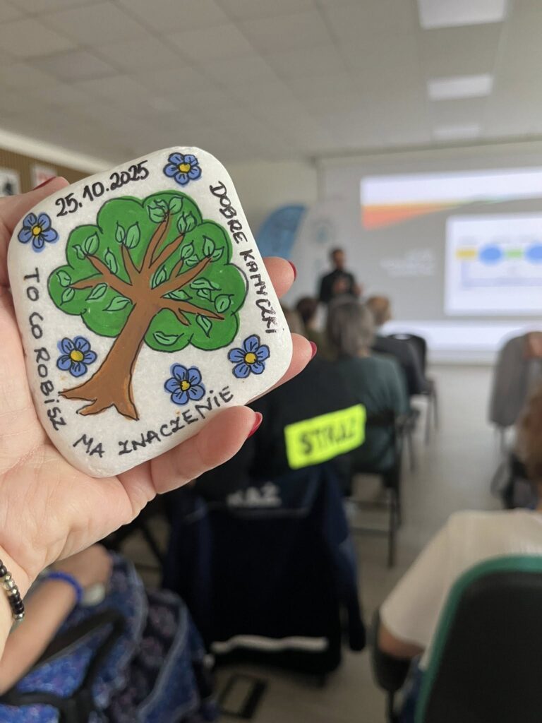 Ręka trzyma pomalowany kamień z drzewem i napisem otoczony kwiatami. W tle widać salę konferencyjną z prezentacją. Kolory to zieleń, błękit i brąz.