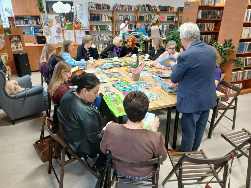 Grupa osób siedzi przy stole w bibliotece, przeglądając książki. Na stole kolorowe książki i roślina. Tło z drewnianymi regałami pełnymi książek.