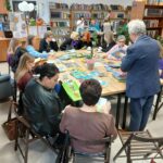 Grupa osób siedzi przy stole w bibliotece, przeglądając książki. Na stole kolorowe książki i roślina. Tło z drewnianymi regałami pełnymi książek.