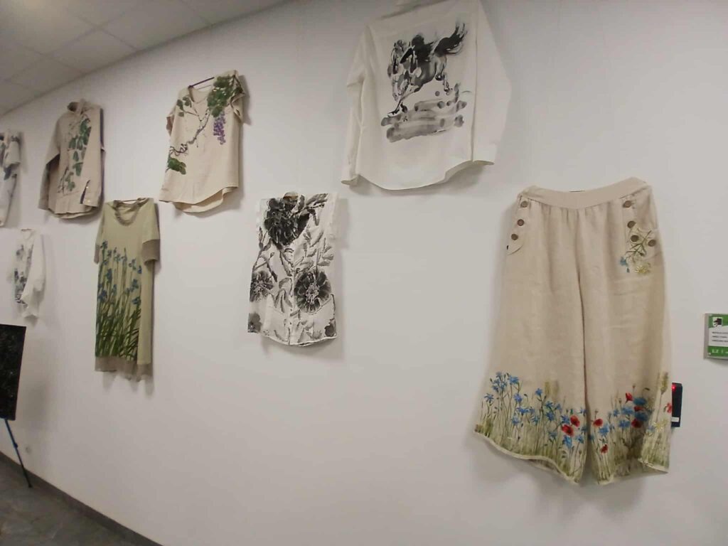 Na białej ścianie wiszą jasno-kremowe ubrania z kwiatowymi i zwierzęcymi wzorami. Tło jest jednolite, co podkreśla szczegóły odzieży. Dominują subtelne kolory natury, jak zieleń i błękit.