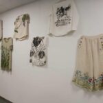 Na białej ścianie wiszą jasno-kremowe ubrania z kwiatowymi i zwierzęcymi wzorami. Tło jest jednolite, co podkreśla szczegóły odzieży. Dominują subtelne kolory natury, jak zieleń i błękit.