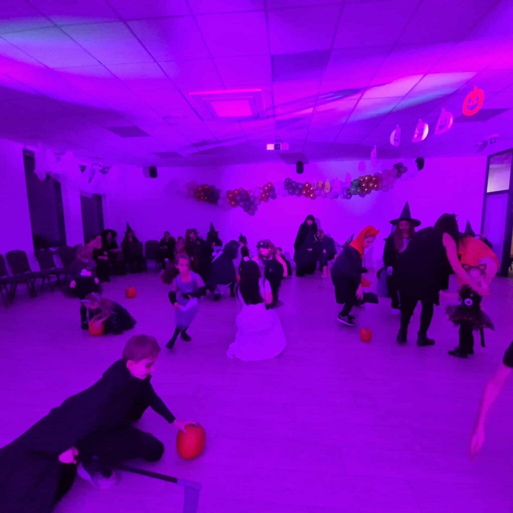 Sala z fioletowym oświetleniem, dzieci w kostiumach na Halloween, dynie na podłodze, kolorowe balony na ścianie.
