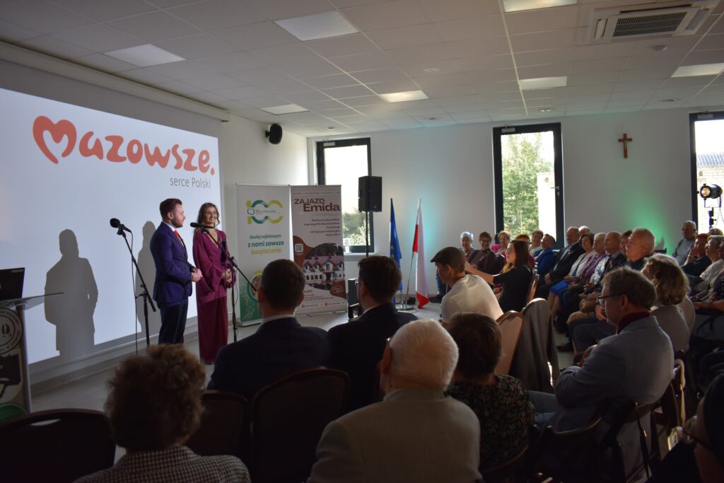 Dwoje ludzi przemawia na scenie przy białym ekranie z napisem Mazowsze. Obok stoją dwa banery. Publiczność siedzi w jasnym pomieszczeniu.