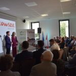 Dwoje ludzi przemawia na scenie przy białym ekranie z napisem Mazowsze. Obok stoją dwa banery. Publiczność siedzi w jasnym pomieszczeniu.