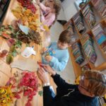 Dzieci i dorosły tworzą jesienne ozdoby z liści przy drewnianym stole. Liście są kolorowe, od czerwonych po żółte. W tle widać półki pełne książek.