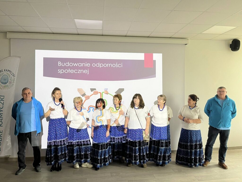 Grupa ludzi stoi przed ekranem z napisem „Budowanie odporności społecznej”. Kobiety ubrane w białe bluzki i niebieskie spódnice. Tło jasne, z dużym ekranem multimedialnym.