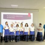 Grupa ludzi stoi przed ekranem z napisem „Budowanie odporności społecznej”. Kobiety ubrane w białe bluzki i niebieskie spódnice. Tło jasne, z dużym ekranem multimedialnym.