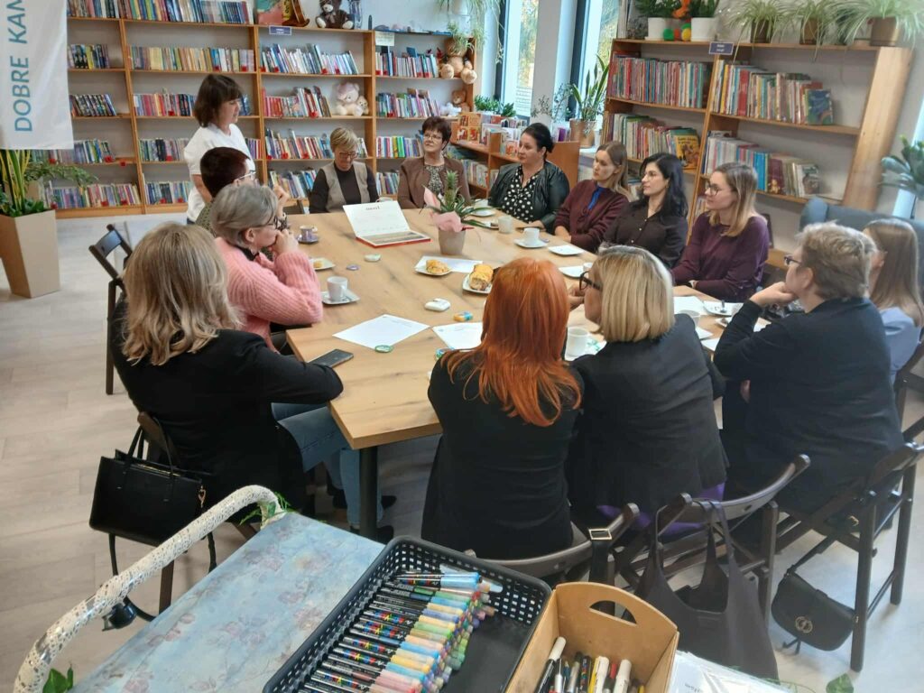 Grupa ludzi siedzi przy drewnianym stole w bibliotece, otoczeni regałami pełnymi kolorowych książek. W tle są rośliny, a na stole kawa i ciasto. Jasne wnętrze z dużymi oknami.