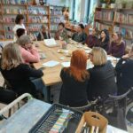 Grupa ludzi siedzi przy drewnianym stole w bibliotece, otoczeni regałami pełnymi kolorowych książek. W tle są rośliny, a na stole kawa i ciasto. Jasne wnętrze z dużymi oknami.