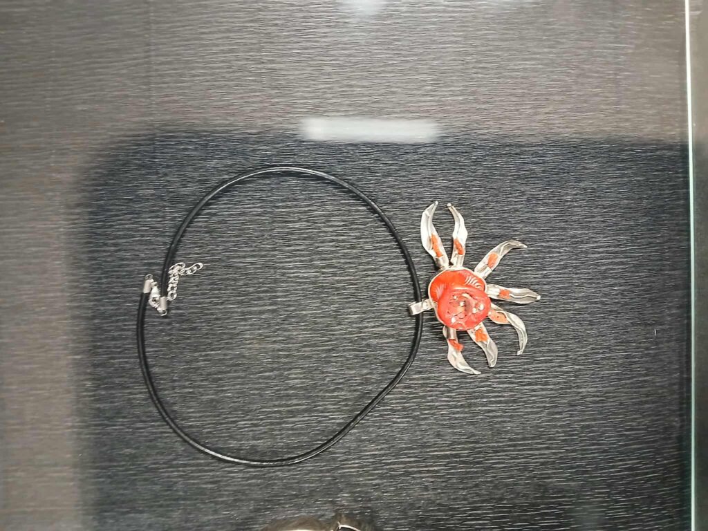 Naszyjnik z czarnym sznurkiem w kształcie kraba na ciemnym tle. Krab jest srebrno-czerwony. Tło ma fakturę szczotkowanego metalu.