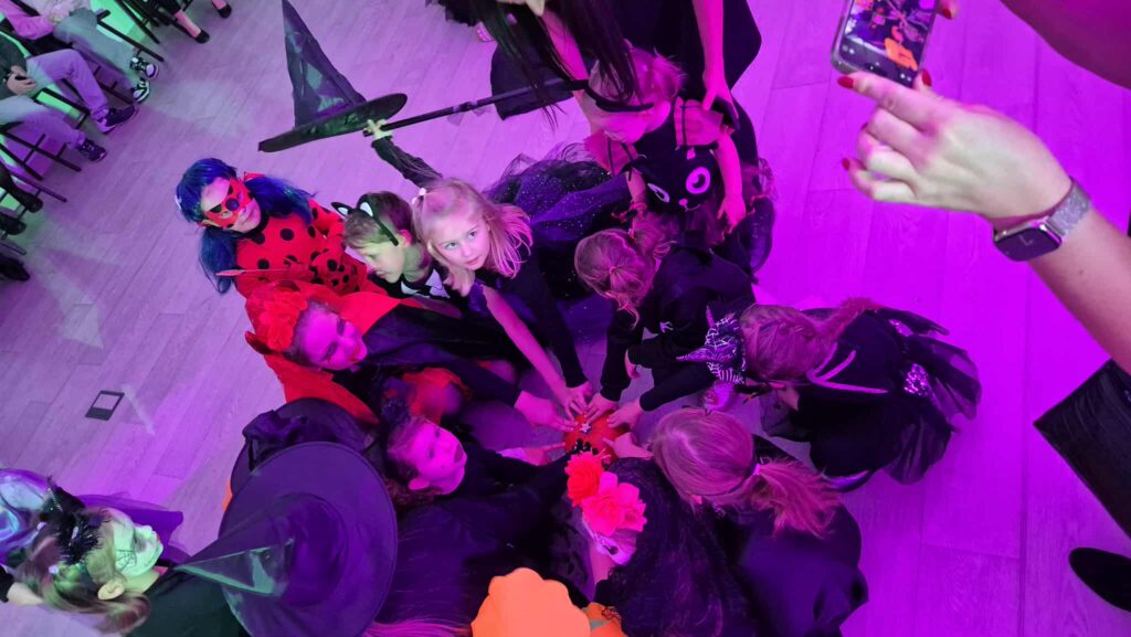 Grupa dzieci w kostiumach halloweenowych siedzi w kręgu, ręce wyciągnięte do środka. Tło jest fioletowe. Na podłodze drewno.