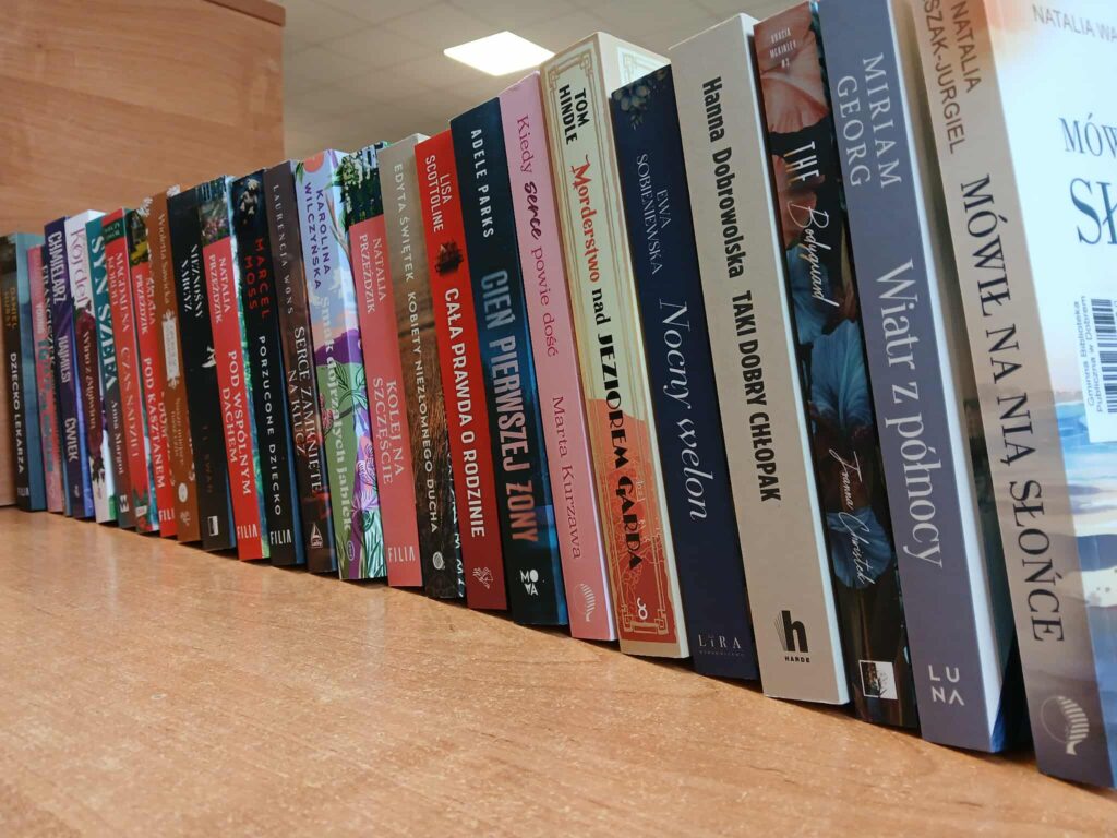 Na zdjęciu widać rząd książek ustawionych pionowo na drewnianym blacie. Okładki są kolorowe, a w tle znajduje się jasna ściana. Oświetlenie jest jasne, prawdopodobnie sztuczne.