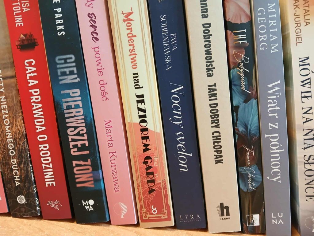 Zdjęcie przedstawia rząd książek na półce, z kolorowymi grzbietami w odcieniach czerwieni, niebieskiego i beżu. Tło jest neutralne, drewniane. Grzbiety zawierają tytuły i nazwiska autorów.