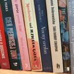 554973294_1325665605778722_8783001144931227119_n Zdjęcie przedstawia rząd książek na półce, z kolorowymi grzbietami w odcieniach czerwieni, niebieskiego i beżu. Tło jest neutralne, drewniane. Grzbiety zawierają tytuły i nazwiska autorów.