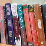 555530640_650980718083749_5104711031300569394_n Półka z kolorowymi książkami; różne tytuły, głównie w pionie. Tło jasne, dominują odcienie czerwieni i fioletu.