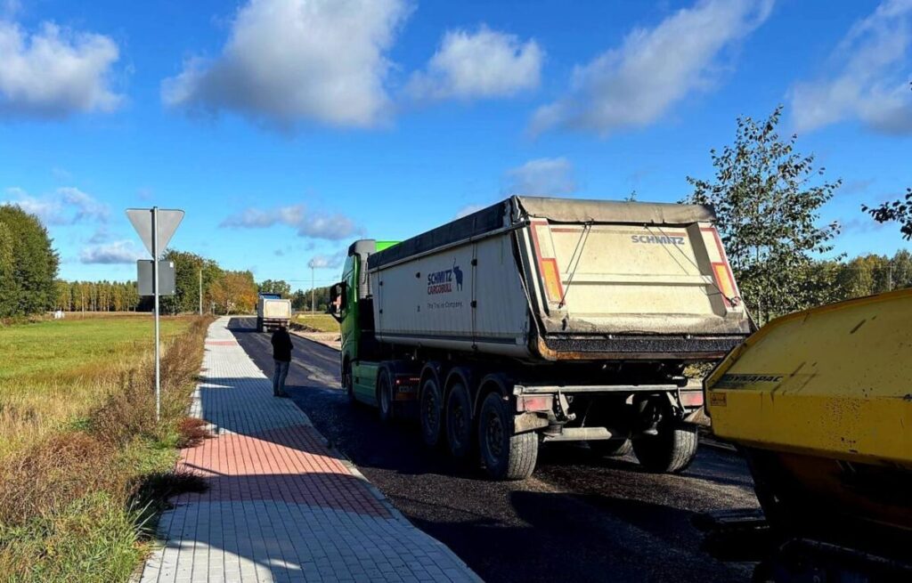 Ciężarówka na asfalcie przy polu, niebieskie niebo z chmurami. Obok chodnik z przechodniem. Drzewa w tle.