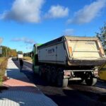 Ciężarówka na asfalcie przy polu, niebieskie niebo z chmurami. Obok chodnik z przechodniem. Drzewa w tle.