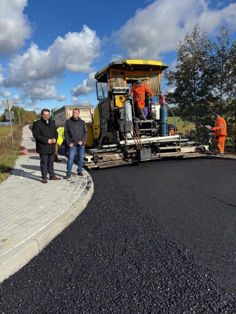 Na obrazie jest maszyna do układania asfaltu otoczona przez kilku pracowników w odzieży odblaskowej. Tło stanowi niebieskie niebo z białymi chmurami oraz drzewa. Na chodniku stoją dwaj mężczyźni w ciemnych kurtkach.