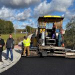 Droga asfaltowana przez maszynę, trzech mężczyzn w ubraniach roboczych, zielone tło z drzewami i niebieskim niebem.