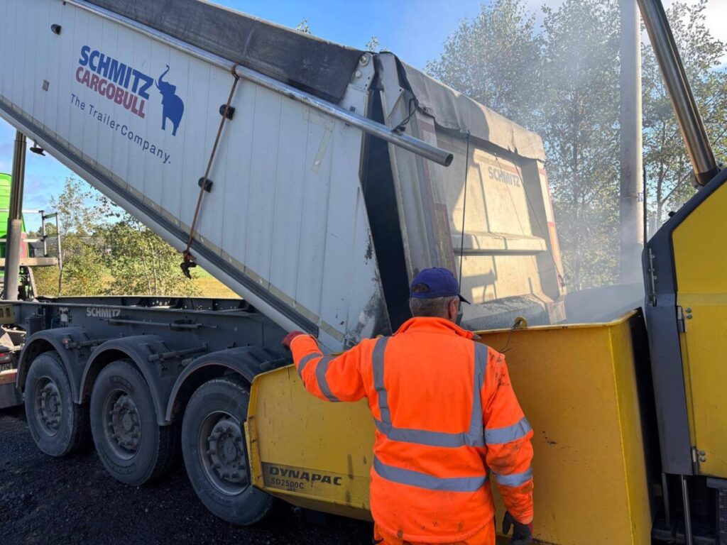 Na zdjęciu widać mężczyznę w pomarańczowym stroju, obok ciężarówki z napisem Schmitz Cargobull. Pojazd ma szare koła, ładunek wysypuje się do żółtej maszyny. W tle jest niebo i zieleń dr