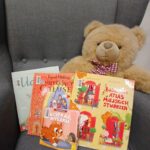 Pluszowy miś siedzi na szarym fotelu obok pięciu książek dla dzieci w kolorowych okładkach. Tło to białe ściany i rozmyte okno. Miś ma czerwoną kokardę w kratkę.