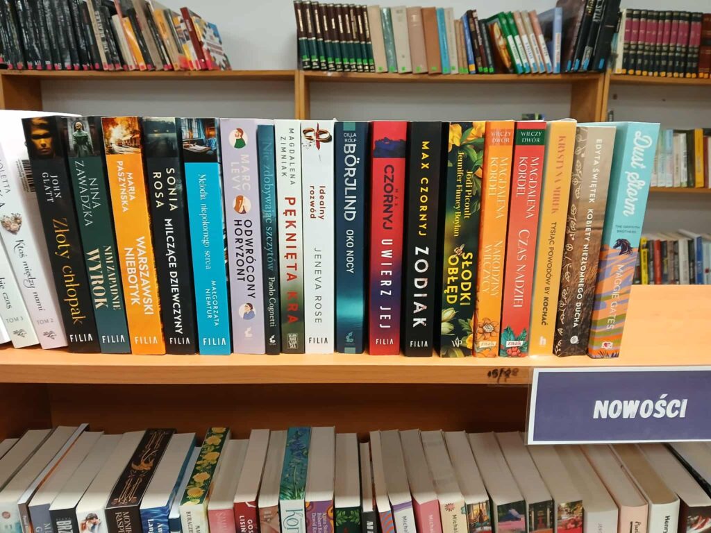 Na zdjęciu widoczna jest półka z książkami w bibliotece. Okładki mają różnorodne kolory i wzory. W tle są inne regały z książkami.