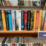 Na zdjęciu widoczna jest półka z książkami w bibliotece. Okładki mają różnorodne kolory i wzory. W tle są inne regały z książkami.