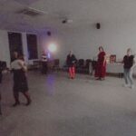 Na zdjęciu widać grupę osób tańczących w jasnym pomieszczeniu. Tło jest białe, z czarnymi oknami i krzyżem na ścianie. Światło odbija się od podłogi, tworząc ciepłą atmosferę.