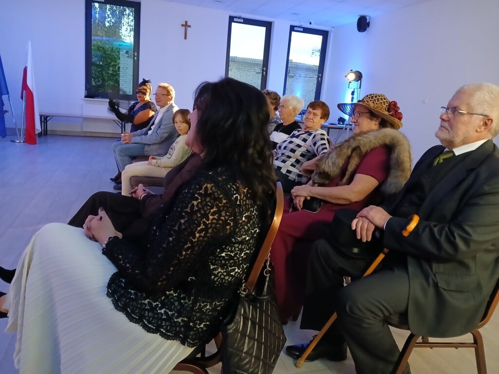 Grupa osób siedzi w jasnym pokoju, tło zdobią okna i krzyż. Niektórzy mają eleganckie stroje, w tym kapelusze. Obok stoi polska flaga.
