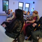 Grupa osób siedzi w jasnym pokoju, tło zdobią okna i krzyż. Niektórzy mają eleganckie stroje, w tym kapelusze. Obok stoi polska flaga.