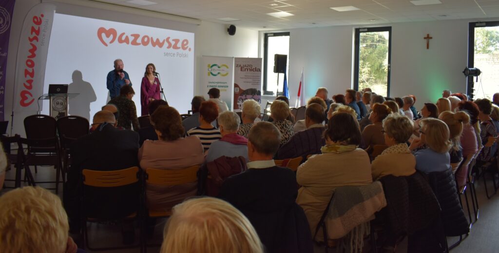 Spotkanie w audytorium, na scenie widoczni są mówca i prezenterka. Jasne tło z logo Mazowsze i szare krzesła. Uczestnicy siedzą tyłem do obiektywu.