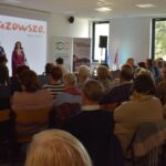 Spotkanie w audytorium, na scenie widoczni są mówca i prezenterka. Jasne tło z logo Mazowsze i szare krzesła. Uczestnicy siedzą tyłem do obiektywu.