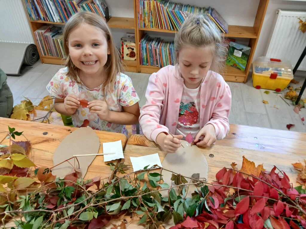 Na zdjęciu dwie dziewczynki przy stole dekorują liśćmi. Stół pokryty jest liśćmi w odcieniach czerwieni i zieleni. W tle półki z książkami.