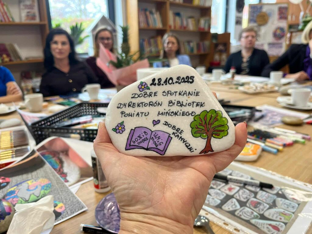 Na zdjęciu widać rękę trzymającą ozdobiony kamień z napisem i rysunkiem drzewa. W tle kobiety siedzą przy stole, na którym są przybory plastyczne. Pomieszczenie ma regały z książkami i jasne ściany.