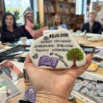 Na zdjęciu widać rękę trzymającą ozdobiony kamień z napisem i rysunkiem drzewa. W tle kobiety siedzą przy stole, na którym są przybory plastyczne. Pomieszczenie ma regały z książkami i jasne ściany.