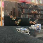 Na zdjęciu widać biżuterię z miedzianymi i srebrnymi elementami z zielonymi i niebieskimi kamieniami, na czarnym tle. W tle półki z książkami w różnych kolorach.