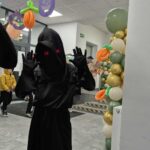 Czarna postać z czerwonymi oczami pozuje w Halloweenowej dekoracji. Tło jest jasne z girlandami dyni. Balony w kolorach złota i zieleni.